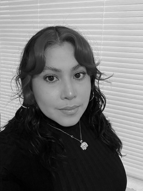 A person with wavy hair and hoop earrings is standing in front of closed blinds, looking at the camera. The image is in black and white. - Elixir Salon | Berkeley, CA