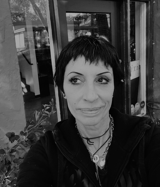 Person with short hair, wearing necklaces and a jacket, takes a selfie outside a building. - Elixir Salon | Berkeley, CA