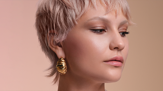 A person with short, platinum blonde hair wears large, gold statement earrings and natural makeup, looking slightly down and to the side against a neutral background. - Elixir Salon | Berkeley, CA