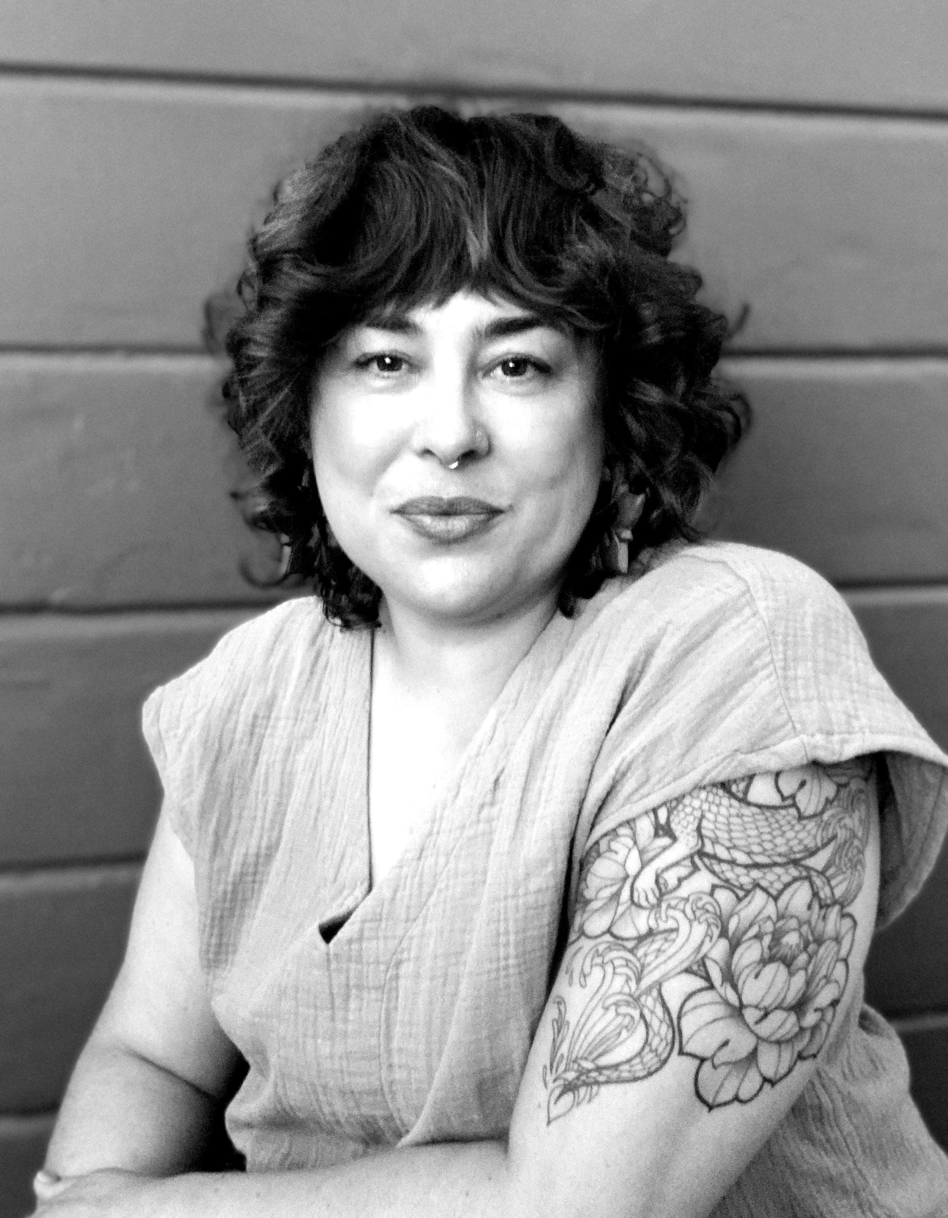 A person with curly hair and a floral tattoo on their arm sits in front of a paneled wall, looking at the camera. - Elixir Salon | Berkeley, CA