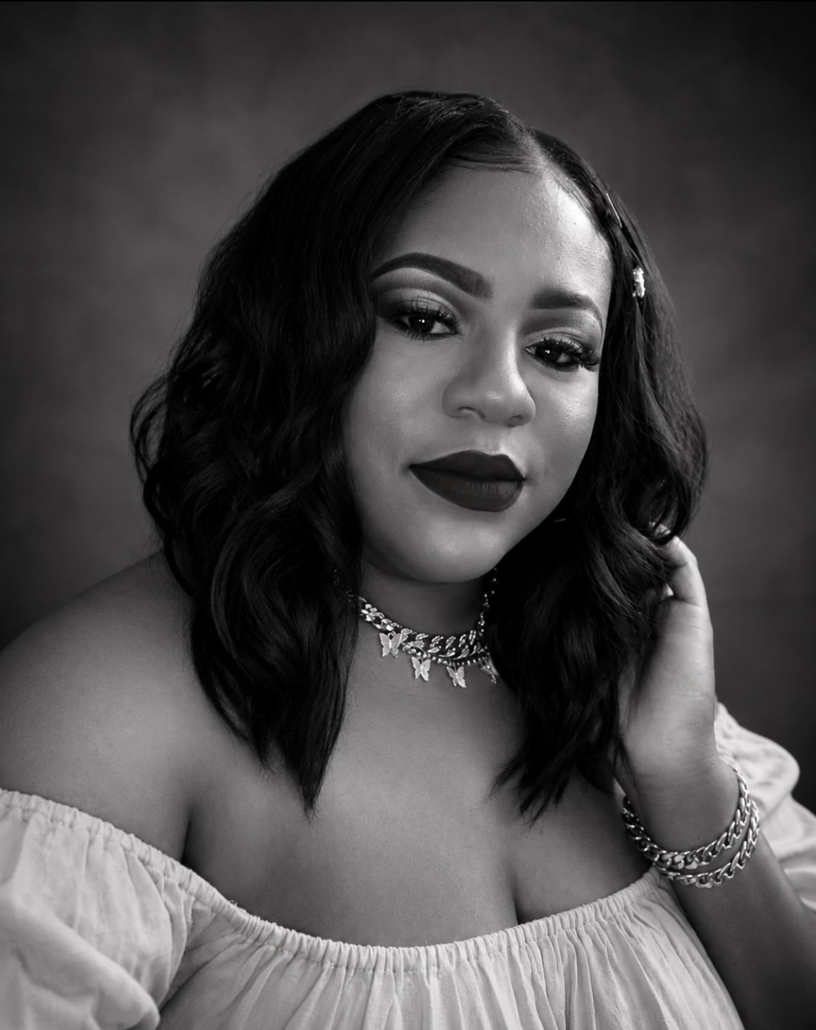 Woman with wavy hair wearing an off-shoulder top, a necklace, and bracelets, posing with one hand near her face against a plain dark background. - Elixir Salon | Berkeley, CA